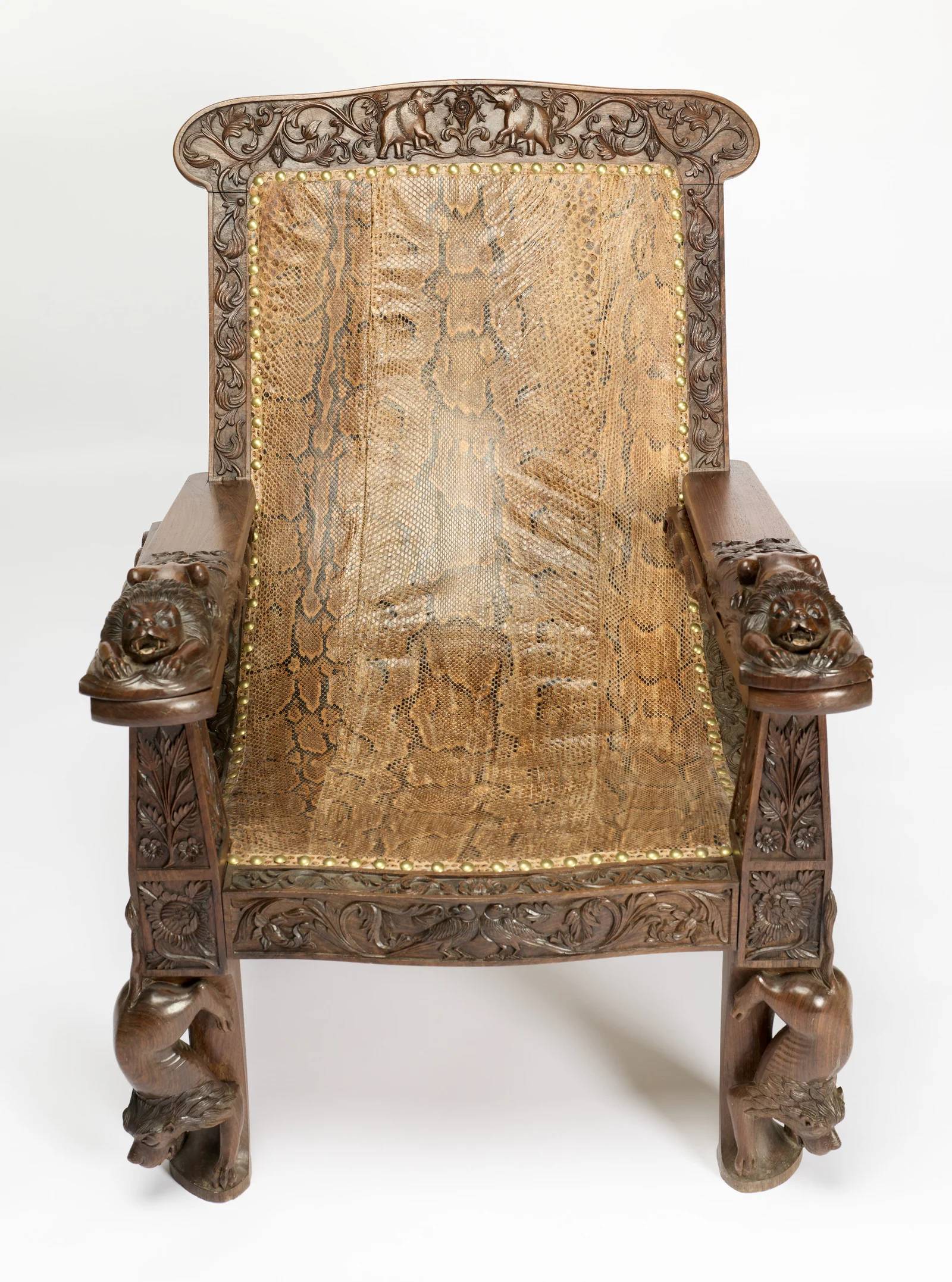 Reptile skin covers the back and seat within a border of scrolling foliage, close-nailed with brass studs, a lion couched at the end of each arm.