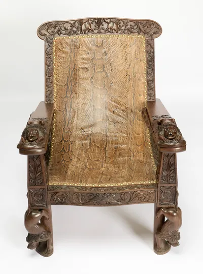 Reptile skin covers the back and seat within a border of scrolling foliage, close-nailed with brass studs, a lion couched at the end of each arm.