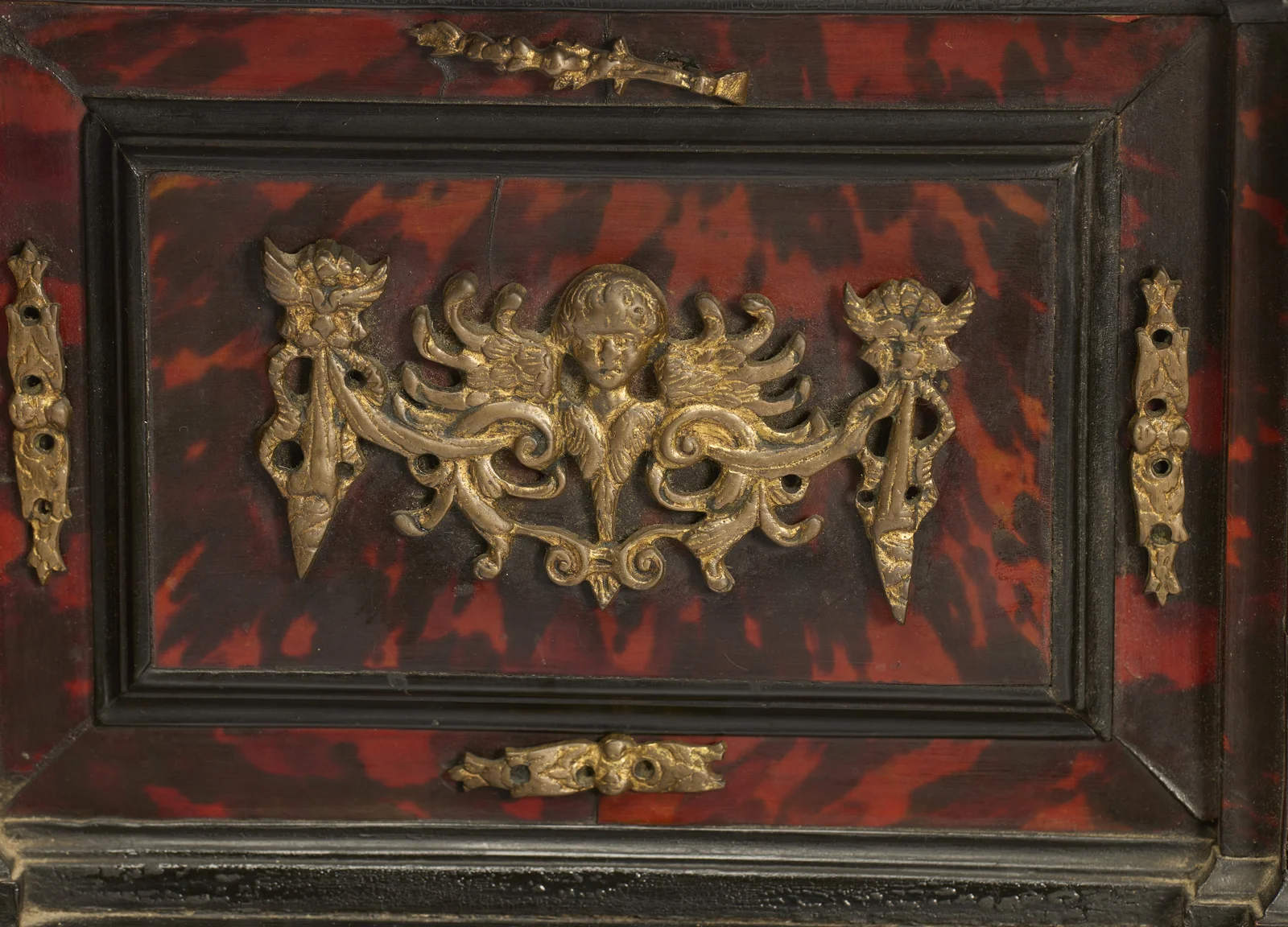 A gilt head between outstretched wings spreads across the mottled red panel of the upper drawer, flanked by pierced mounts with empty fixing holes.