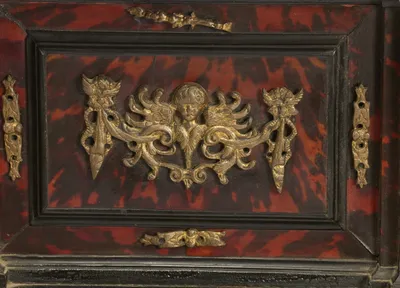A gilt head between outstretched wings spreads across the mottled red panel of the upper drawer, flanked by pierced mounts with empty fixing holes.