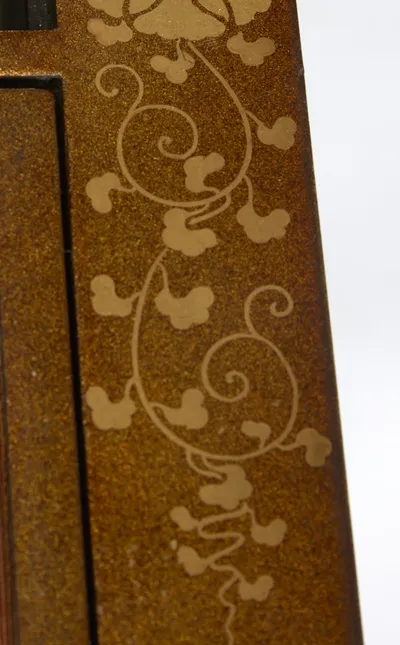 The key-fret border of the fabric meeting gilt peony scrolls on the surround, open wells behind, with wear visible along the door edge above.
