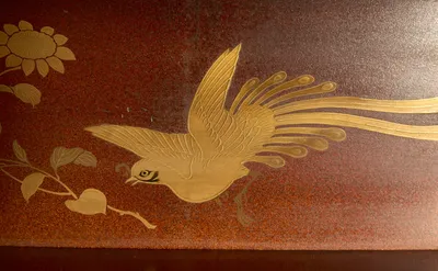 A bird in flight trailing long tail plumes, drawn in gold on the red speckled ground beside a flowering spray.