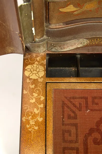 An engraved strap hinge beside the serpentine drawer, trailing gilt scrolls along the border, the lacquer rubbed on the door edge.