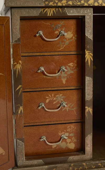 Small drawers with silvered swan-neck handles and flower sprays on a glittering orange-gold ground, framed by engraved mounts and bamboo.