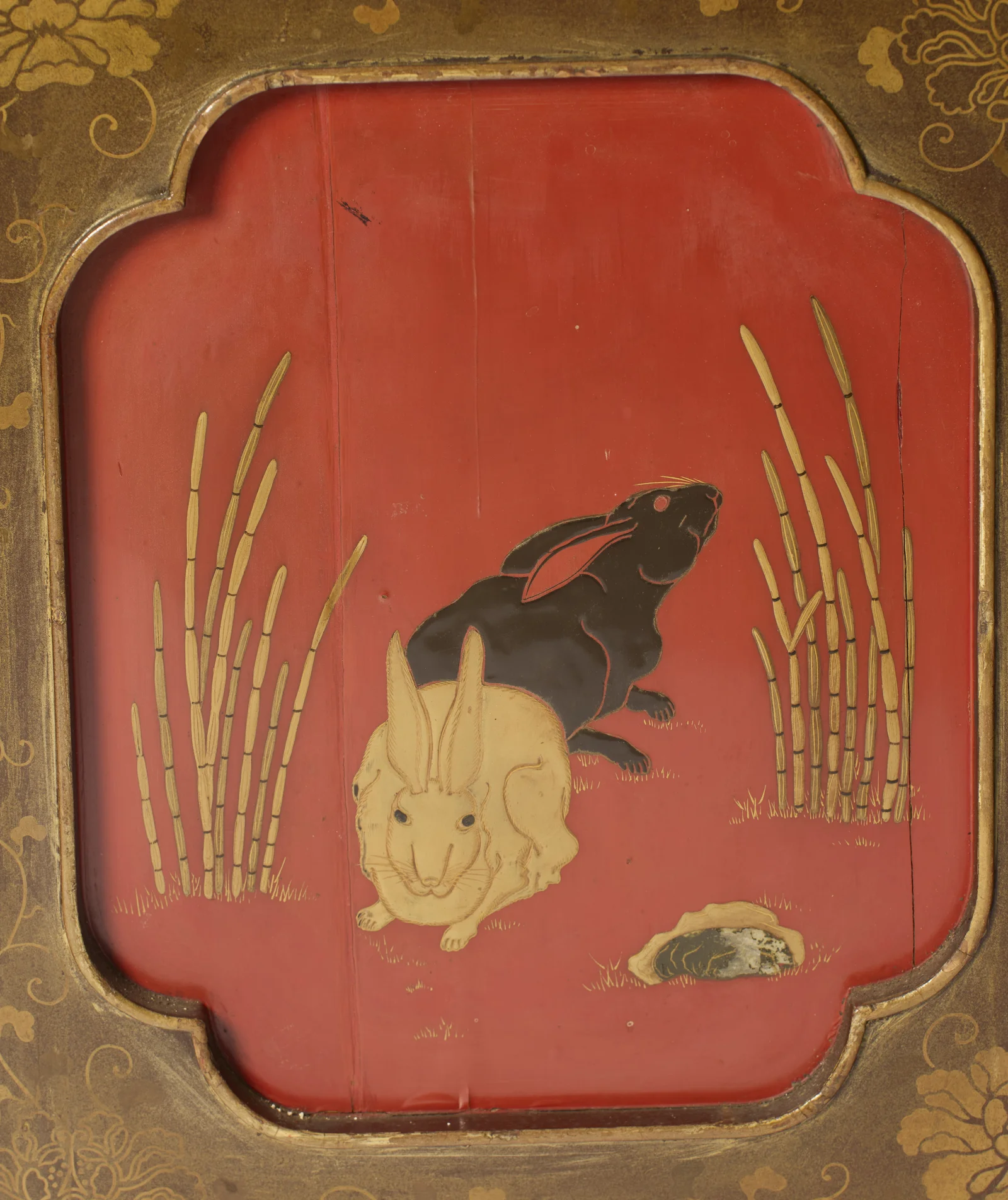 A dark rabbit and a pale rabbit between golden reeds on the red lacquer ground of a shaped side panel.