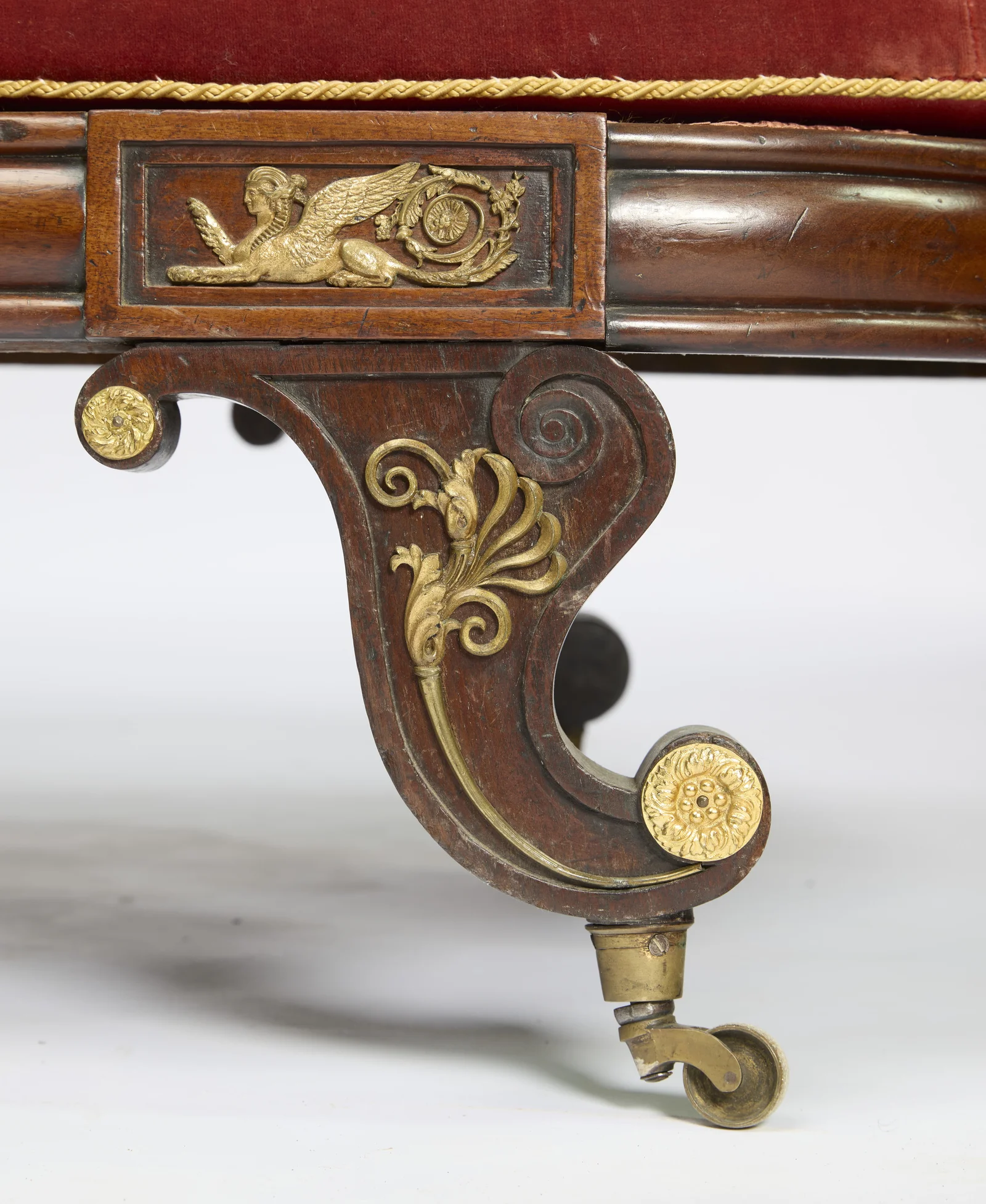 A gilt palmette spray runs down the carved scroll of the leg to a rosette boss and brass castor, beneath a second winged-figure panel on the rail.