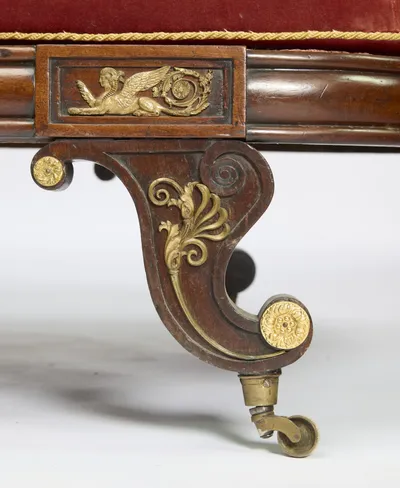A gilt palmette spray runs down the carved scroll of the leg to a rosette boss and brass castor, beneath a second winged-figure panel on the rail.