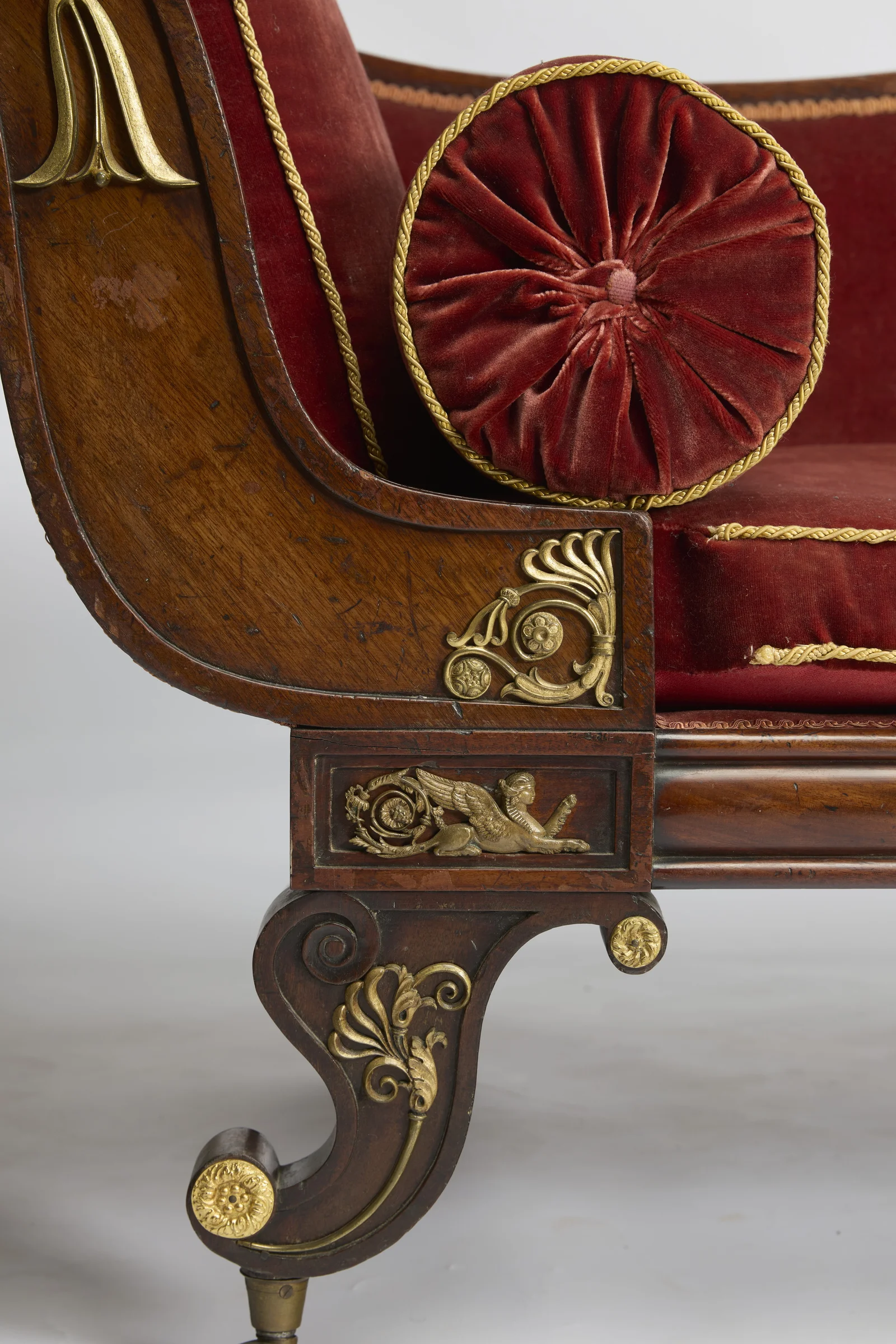 A gilt husk drop falls along the scroll above the round pleated end of the bolster, with a pierced gilt palmette at the angle and a winged-figure panel over the leg.