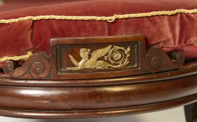A gilt-metal recumbent winged figure beside a wheel wreathed in foliage, applied to a raised panel between carved C-scrolls; a loose loop of the gold cord trim stands proud of the cushion above.