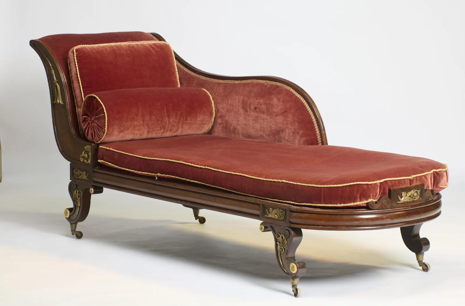 The scrolled end rises above a shaped back that sweeps down towards the foot, the rose velvet squab, pillow and bolster trimmed with gold cord, on scrolled legs with gilt mounts and brass castors.