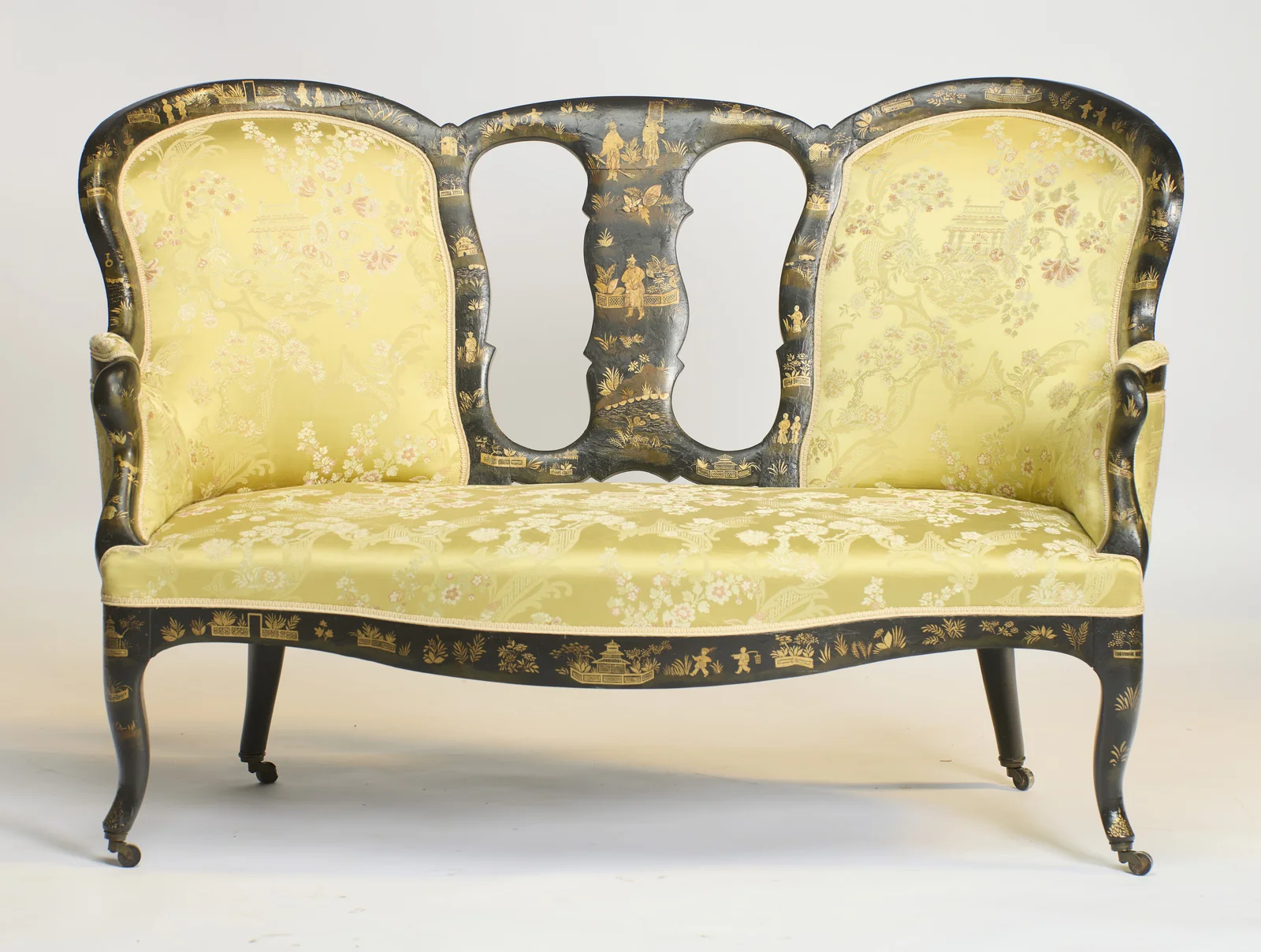 Arched back panels of unequal height flank the pierced centre splat, painted in gilt with figures and pavilions, above a serpentine seat rail and swept legs on castors.