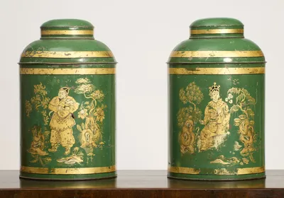 Both canisters face on, each painted in gilt with a chinoiserie figure among trees, flowering plants and rockwork, the left a standing figure holding an animal, the right a seated figure wearing a headdress.