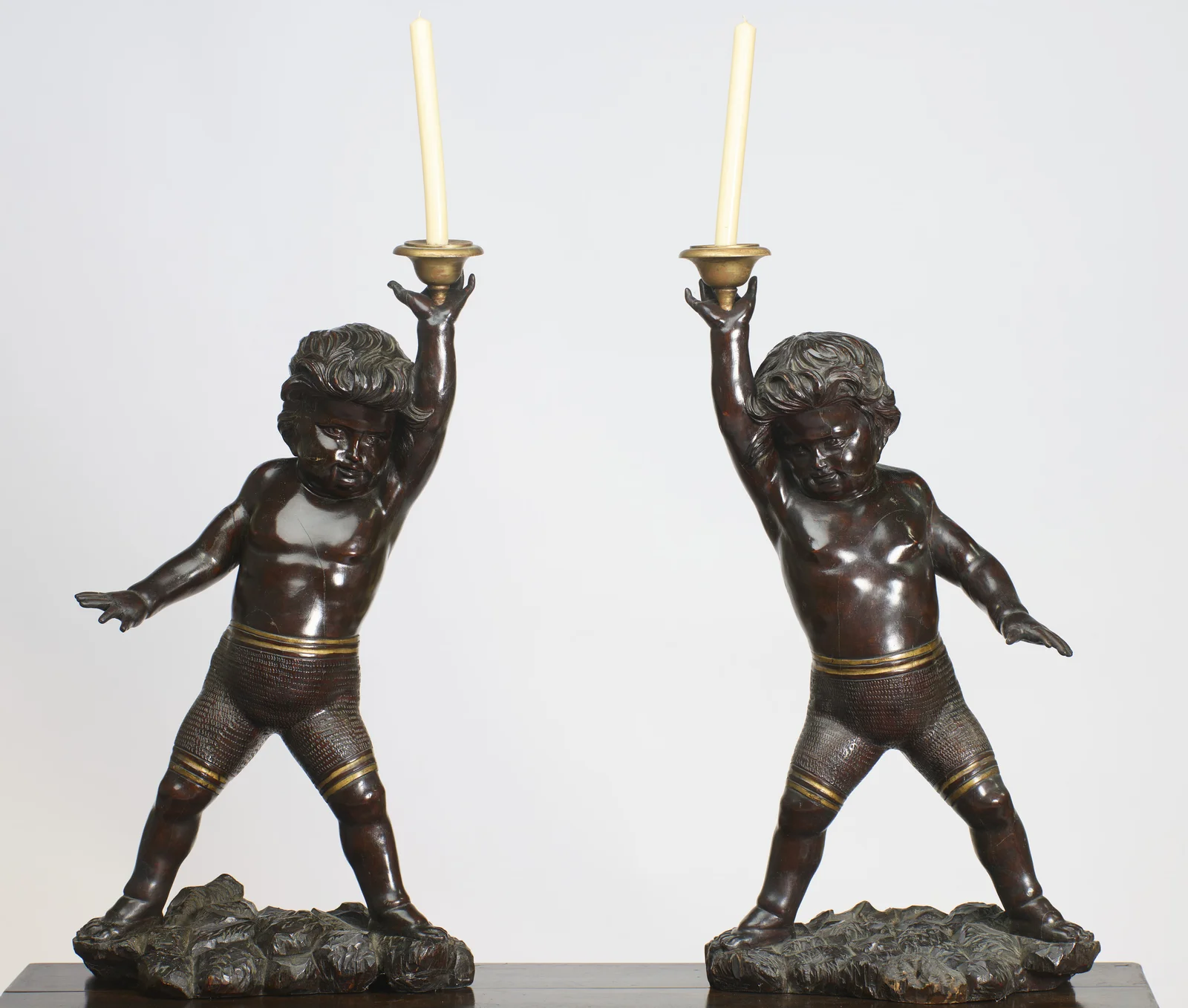 Both figures stride across carved rockwork bases in mirrored poses, each raising a gilt-metal drip-pan and nozzle overhead on an open palm.