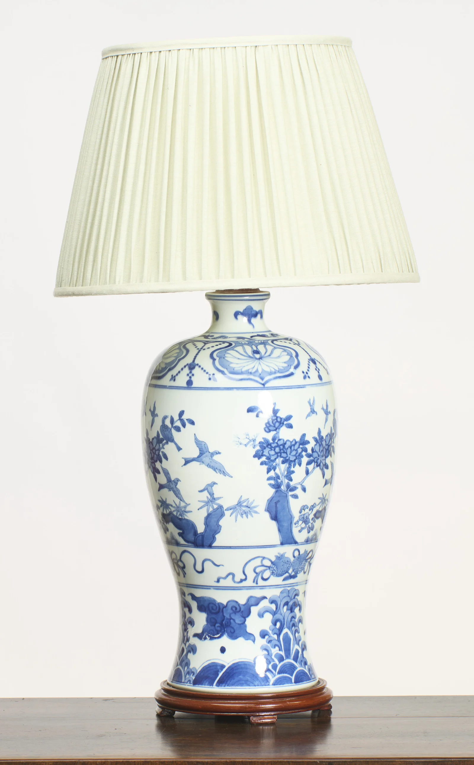 The whole lamp seen straight on, birds in flight among flowering branches across the body, raised on a turned wood base beneath a pleated cream shade.