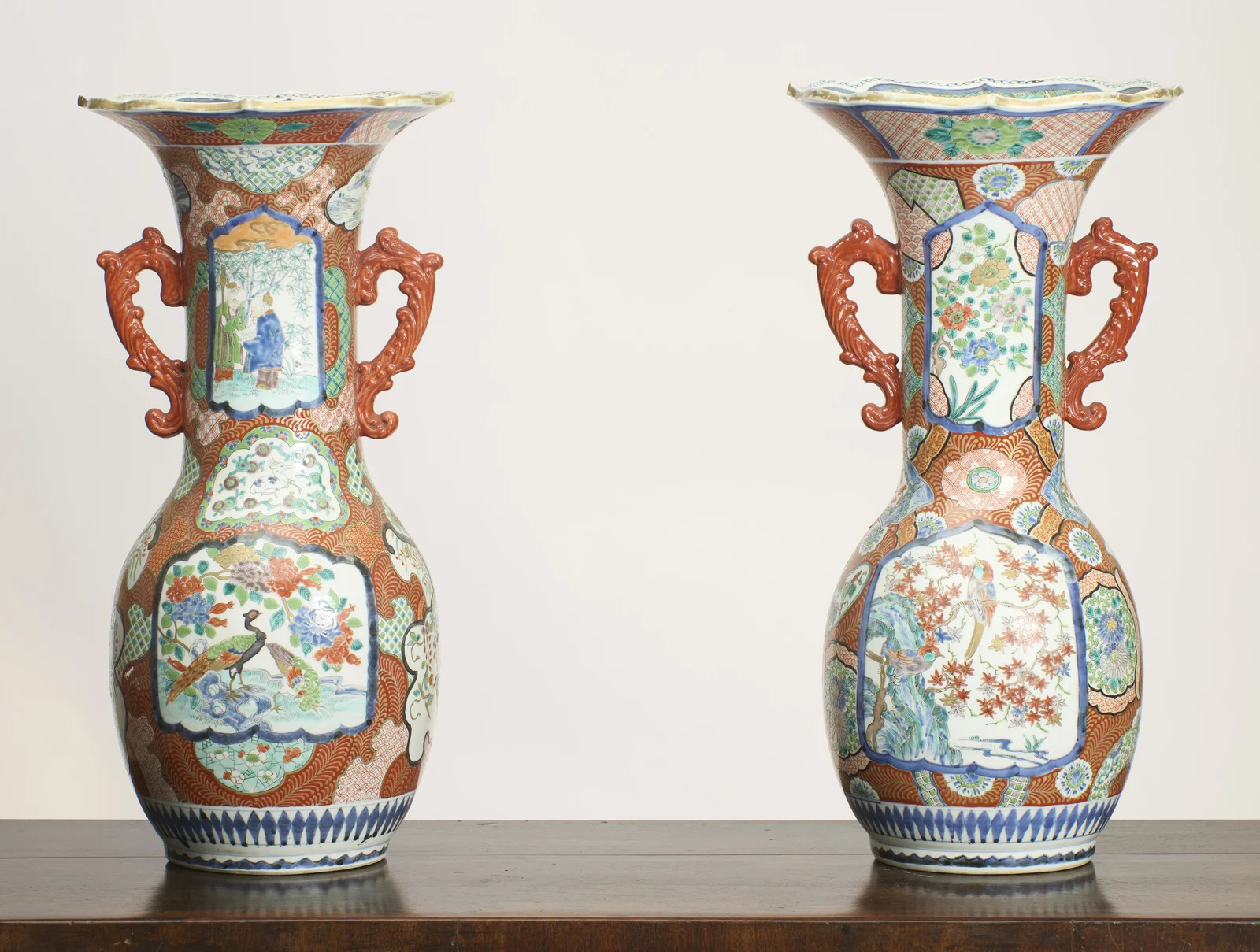 Both vases together, trumpet necks with scalloped gilt-edged rims, coral-red scroll handles, and panelled decoration reserved on an iron-red ground.