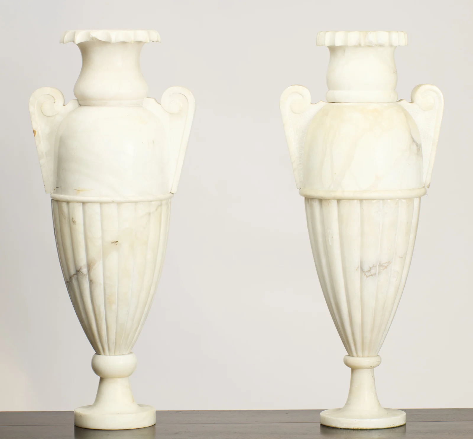 Twin scroll handles rise from domed shoulders above tapering fluted bodies, each urn raised on a knopped stem and circular foot in softly veined white stone.