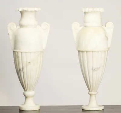 Twin scroll handles rise from domed shoulders above tapering fluted bodies, each urn raised on a knopped stem and circular foot in softly veined white stone.