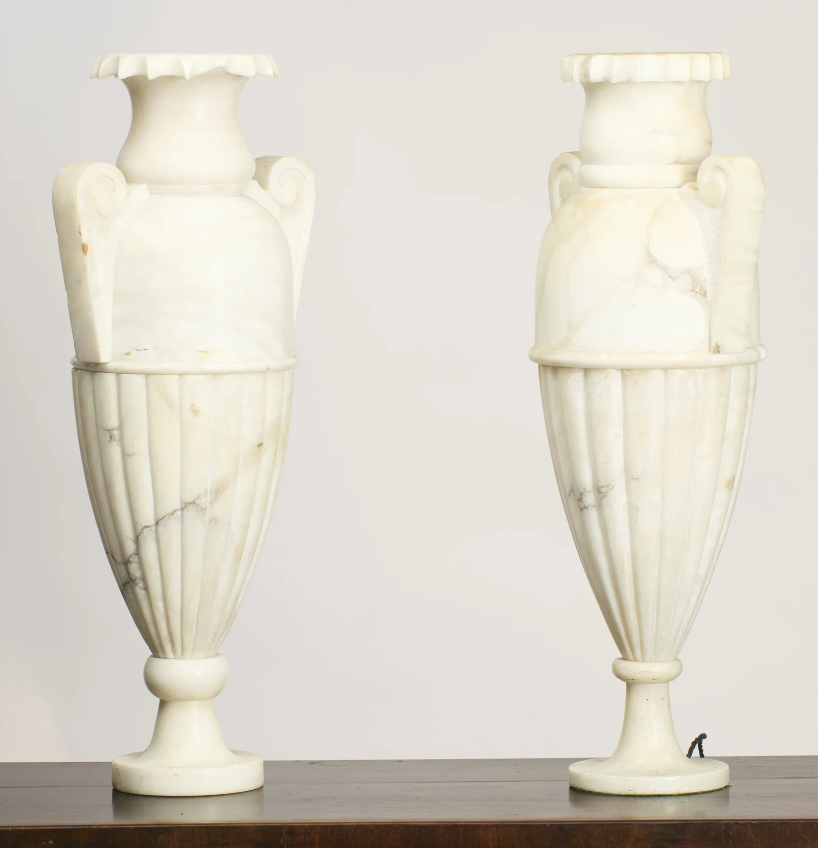 Both urns stand full height on a dark wooden surface, the crimped and fluted rims above the scroll handles, with a braided flex just visible at the right-hand foot.