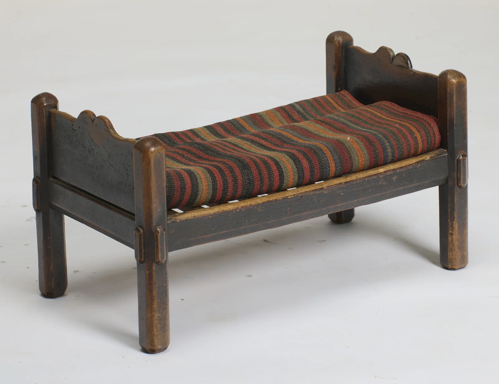 The bed form seen from an angle, shaped end boards between rounded posts, the slatted platform carrying the striped flat-woven cushion, with the dark finish worn to lighter timber on posts and rails.