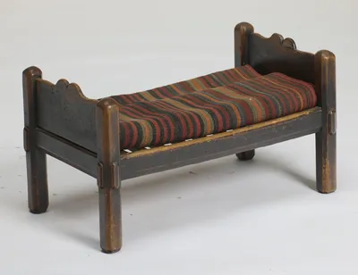 The bed form seen from an angle, shaped end boards between rounded posts, the slatted platform carrying the striped flat-woven cushion, with the dark finish worn to lighter timber on posts and rails.