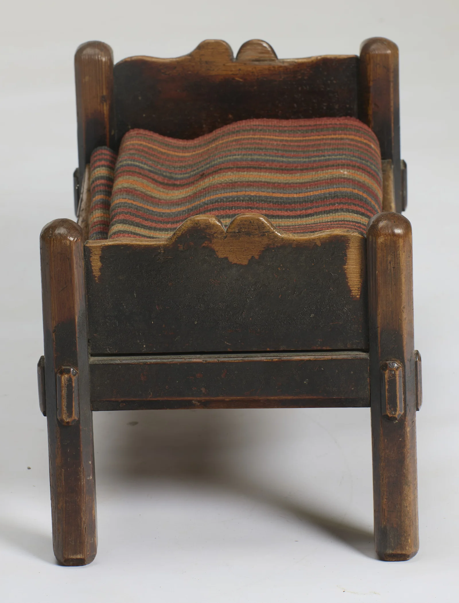 The shaped end board with its double-arched crest between rounded posts, the dark surface rubbed to bare timber along the edges and legs, the striped cushion beyond.