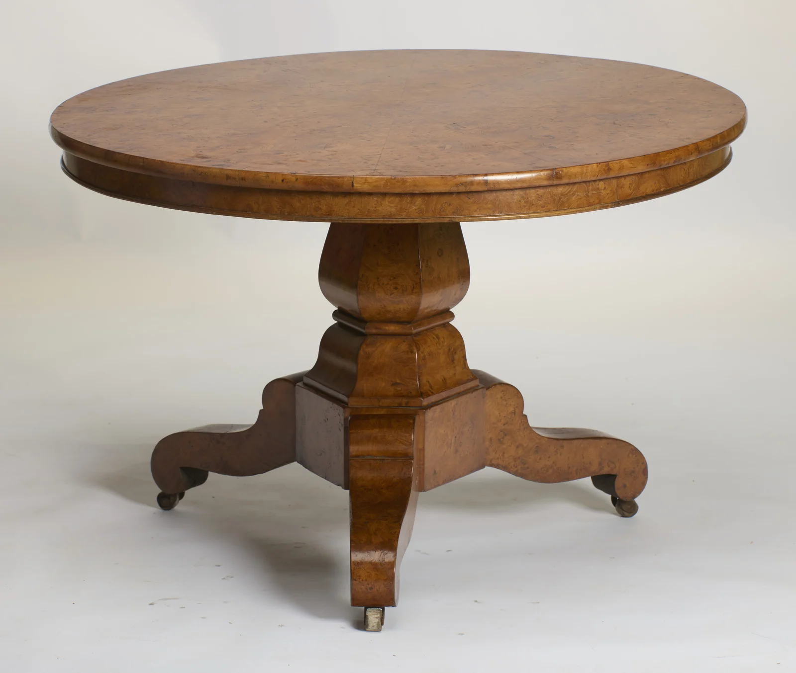 Circular top with dense burr figuring, raised on a faceted baluster pedestal with three scroll legs ending in castors.