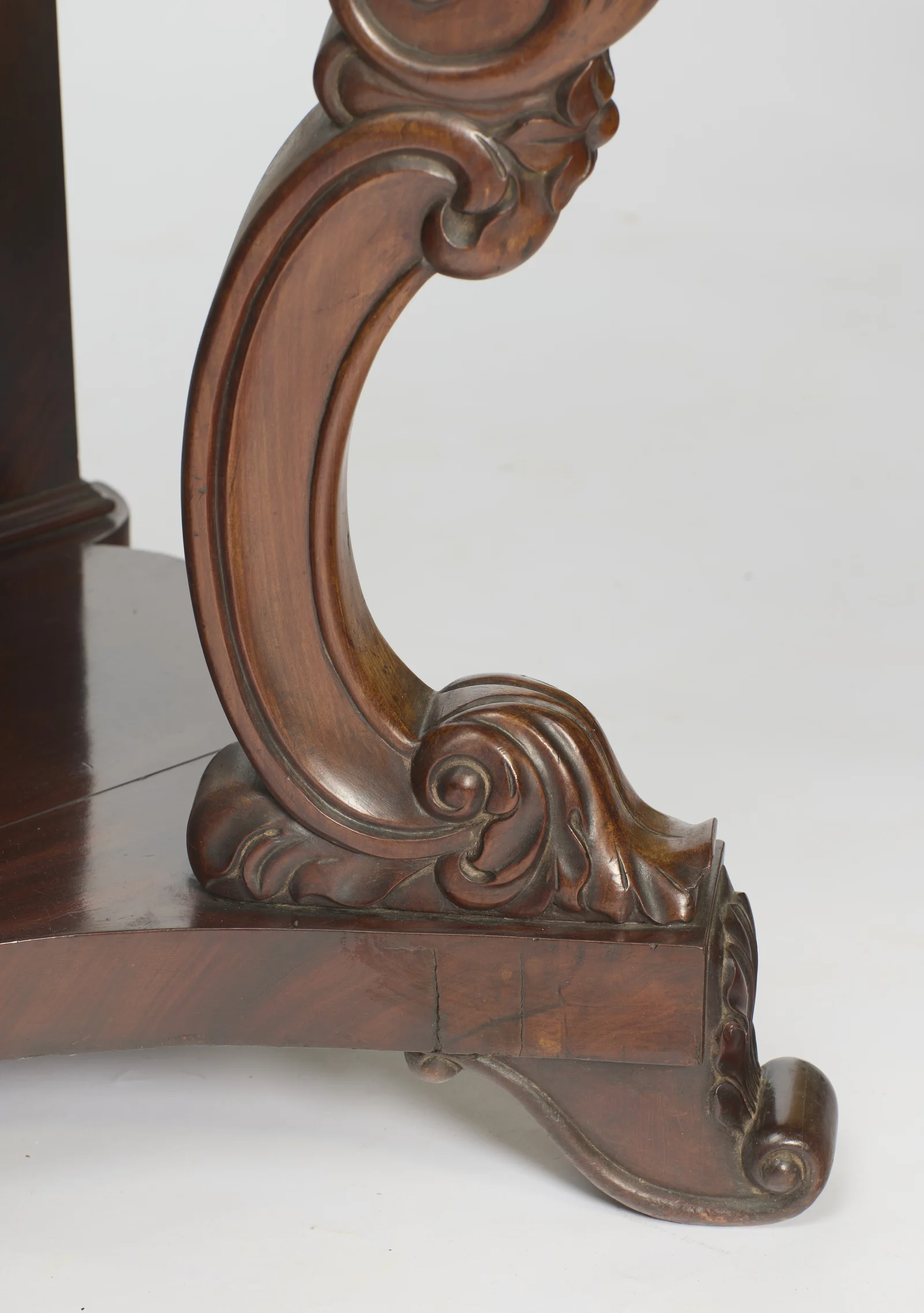 The scroll of the support ends in an acanthus-wrapped foot beside a scrolled toe on the platform base, with old cracks visible on the plinth face below.