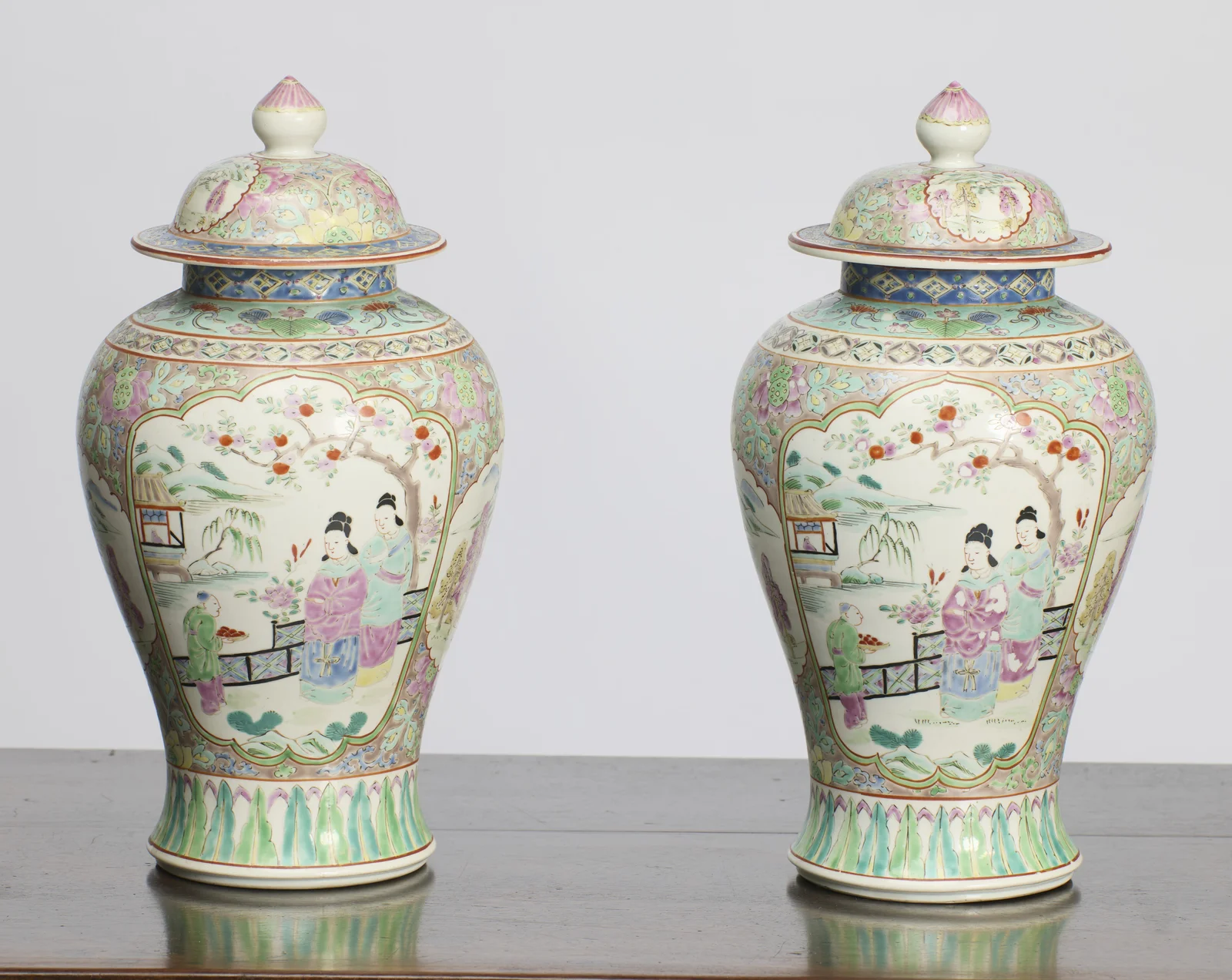 Both vases and covers, each with a shaped panel of two ladies and a boy carrying a tray in a fenced garden beneath a fruiting blossom tree, a pavilion and willow at the water's edge beyond.