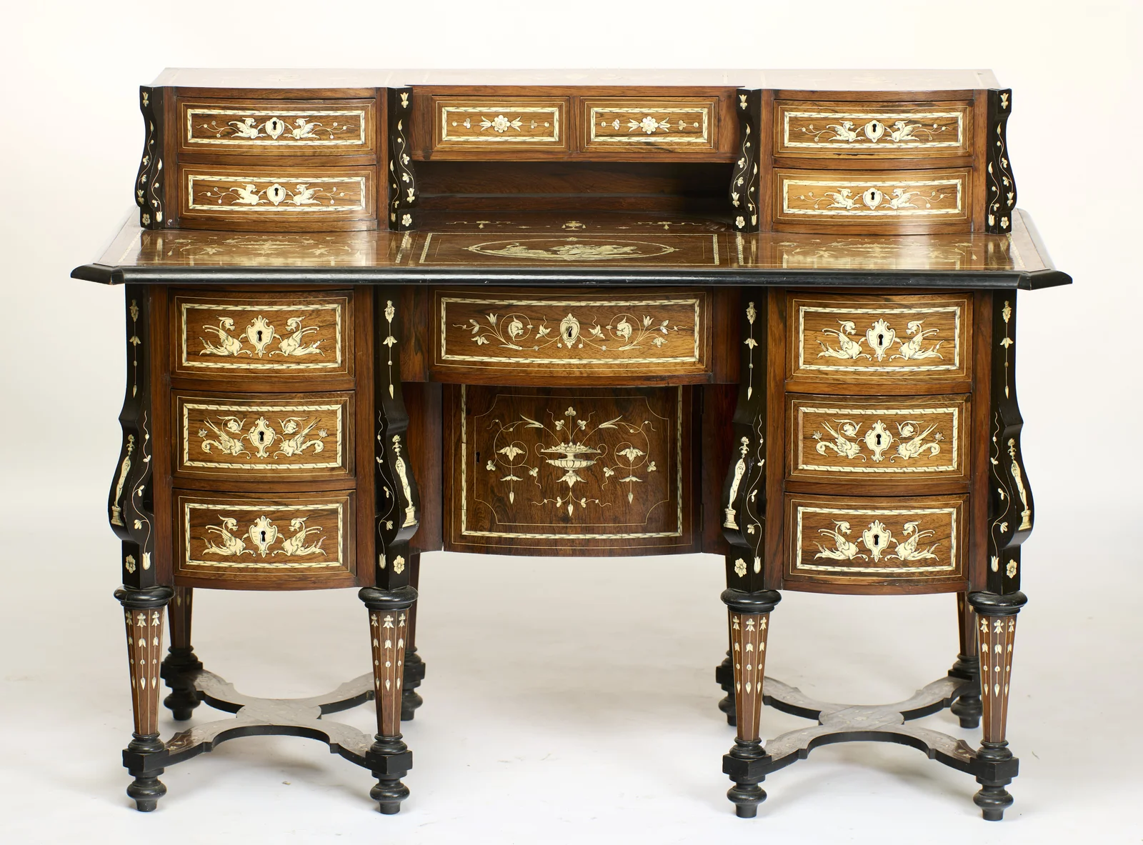 The desk complete: a raised superstructure of small bow-fronted drawers above a frieze drawer and two pedestals of three drawers each, every surface carrying pale engraved inlay within ebonised mouldings, on inlaid tapering legs joined by shaped stre