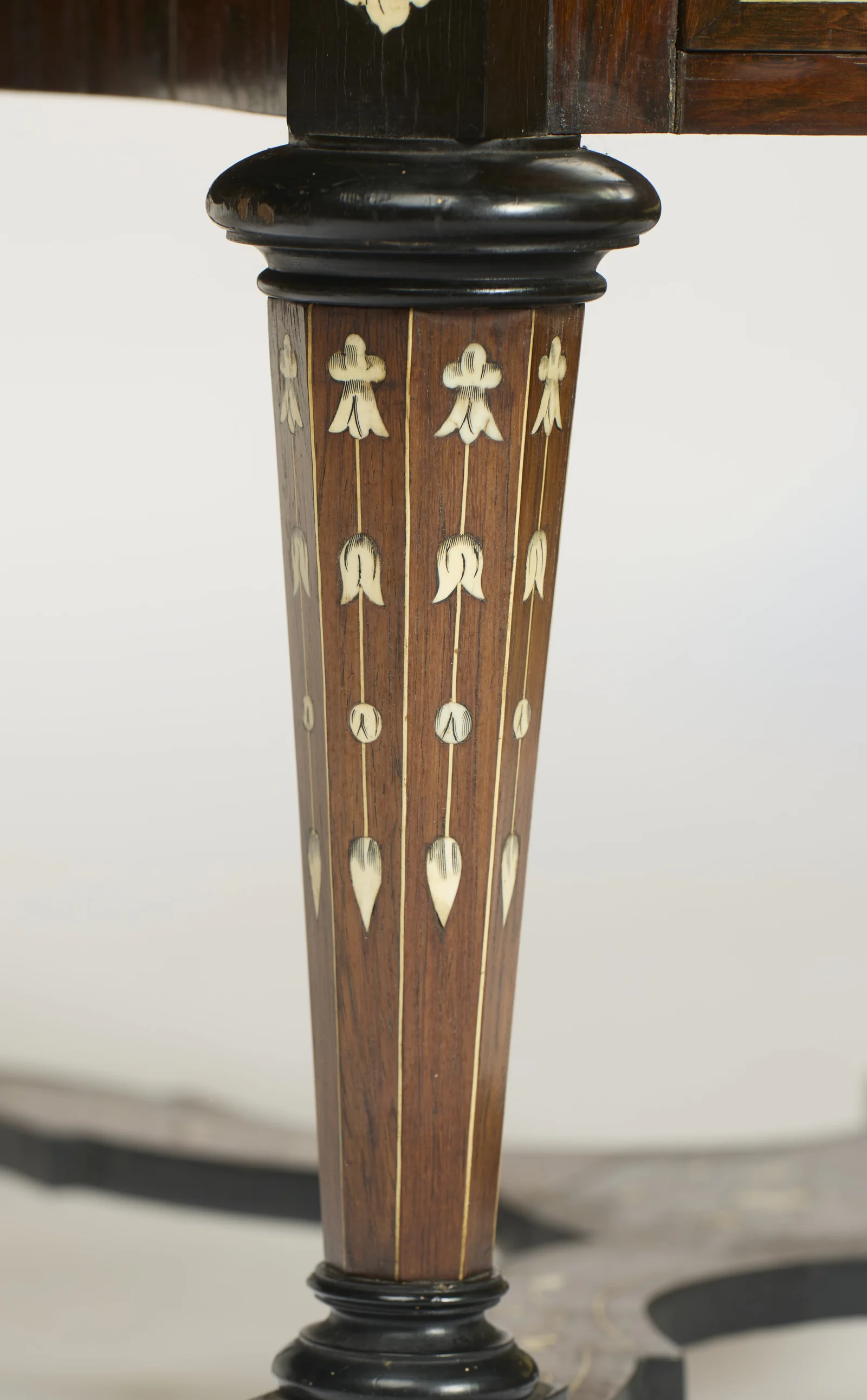 A tapering leg inlaid with pendant lines of husks and tulips between ebonised collars, ending in a turned foot where the shaped X-stretcher meets its block.
