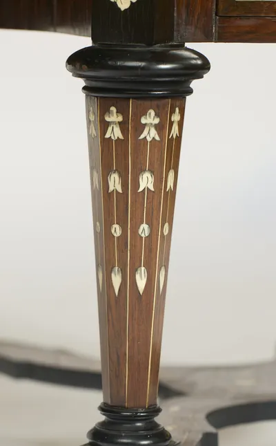 A tapering leg inlaid with pendant lines of husks and tulips between ebonised collars, ending in a turned foot where the shaped X-stretcher meets its block.