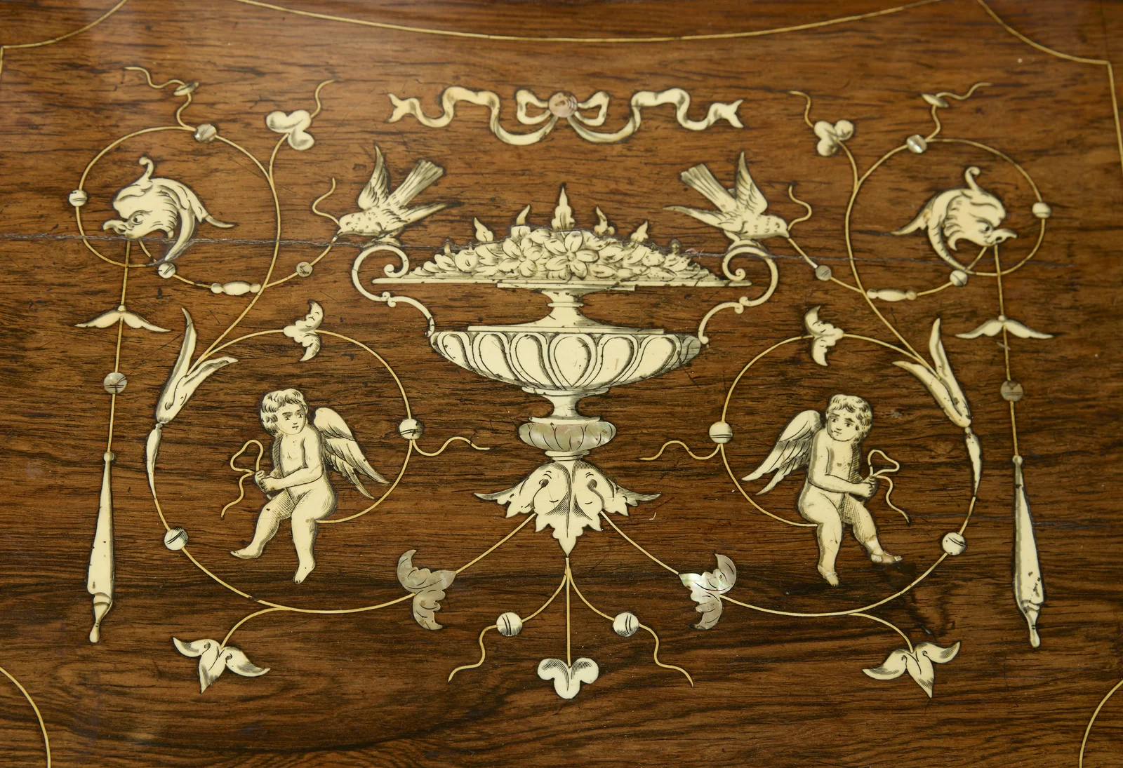 The matching composition on the companion panel of the top: cherubs, birds and dolphin-headed scrolls about a flower-filled urn, with a bow-fronted superstructure drawer above.