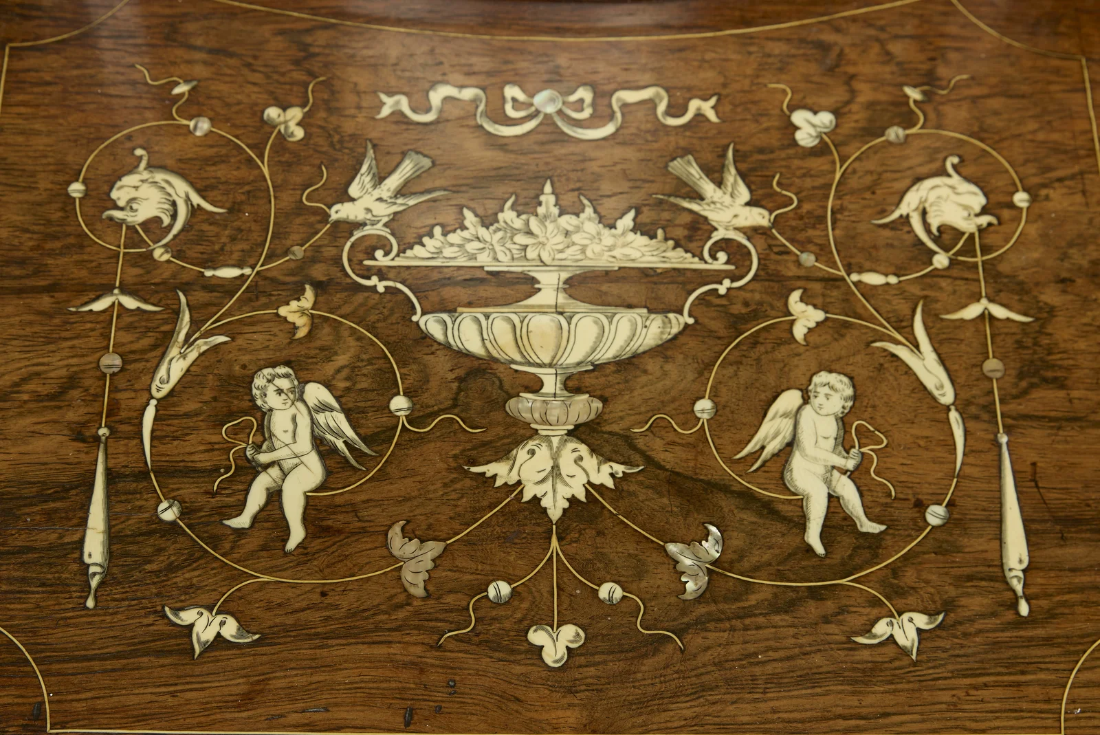 Two winged cherubs holding bows flank a pedestal urn piled with flowers, with songbirds, dolphin-headed scrolls, a ribbon bow above and iridescent leaf accents, all within engraved banding.
