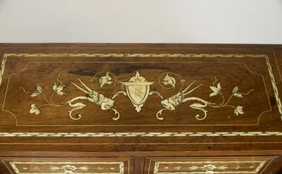 Winged dragon-birds flanking a hatched shield beneath a leafy canopy, with scrolling tulip stems and iridescent details to either side, within engraved chevron banding.