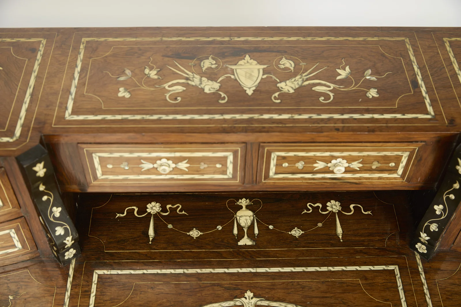 The two central small drawers with flower knobs above the open recess, its back panel inlaid with an urn hung between ribbon-tied swags; the top inlaid with winged creatures about a shield.