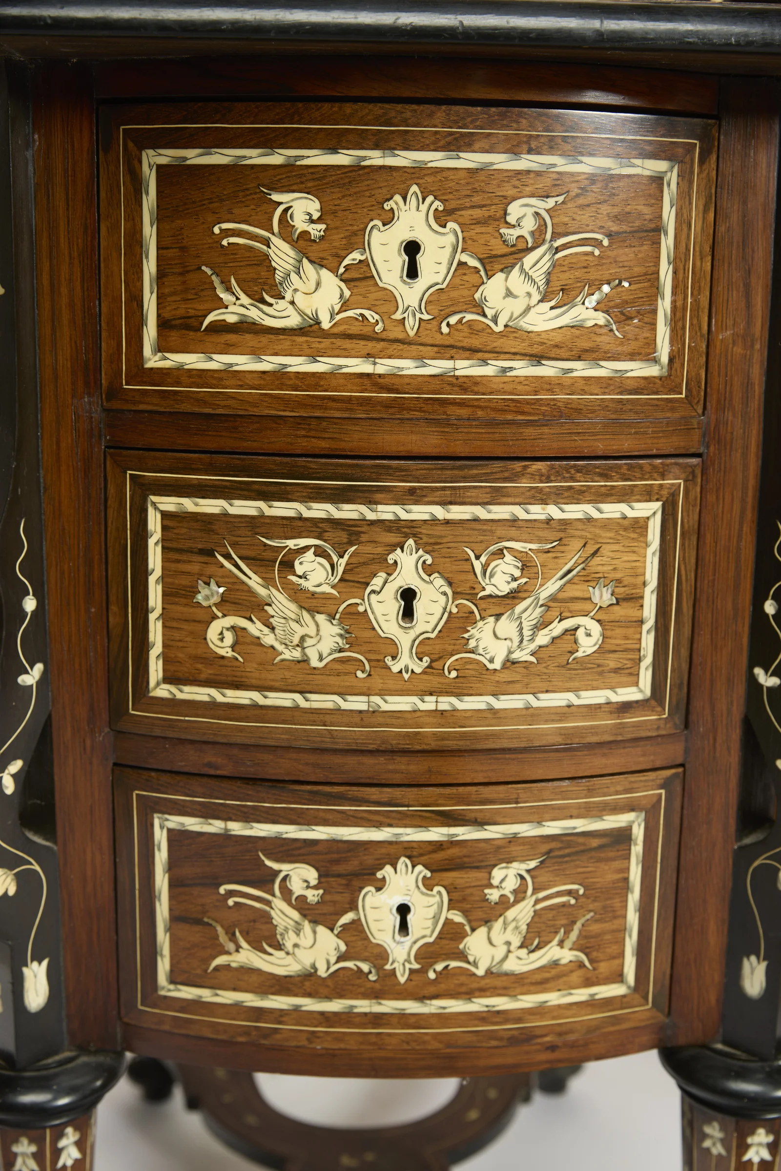 Three bow-fronted drawers, each panel with a pair of winged dragons about a shaped keyhole escutcheon within ribbon-twist banding, the ebonised scroll supports to either side.
