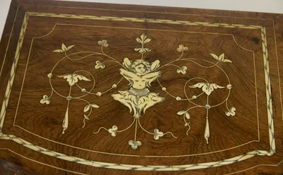 The top of one drawer-box inlaid with a winged half-figure amid looping tendrils, trefoils and drop pendants, with small iridescent accents at the crossings.