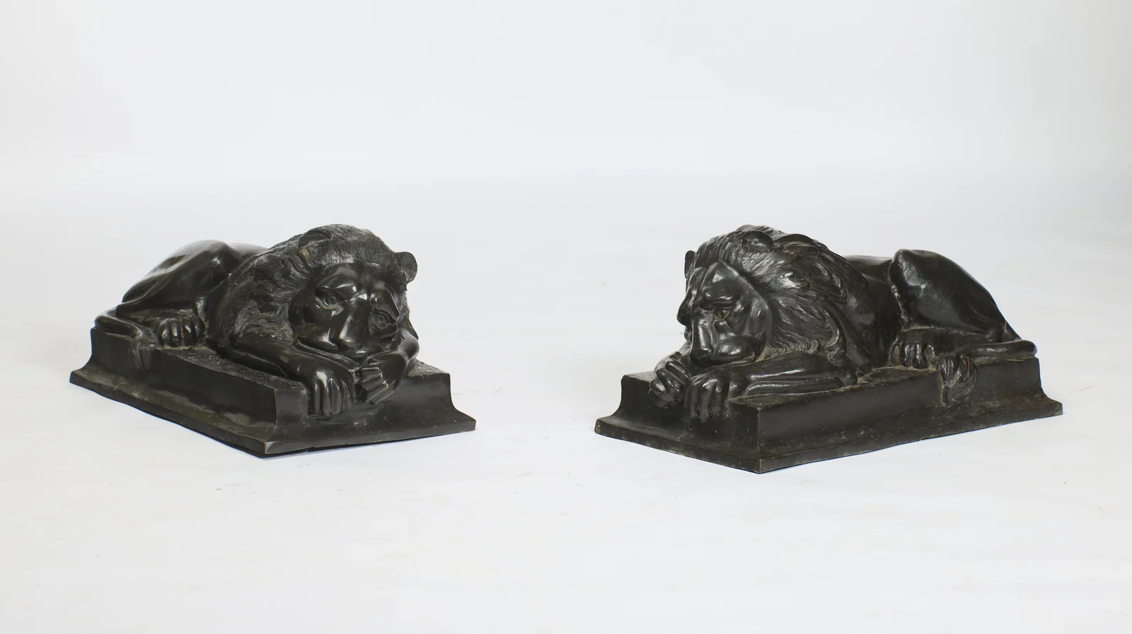 Both lions recumbent and asleep, each head sunk onto crossed forepaws, on rectangular plinths with moulded stepped edges.