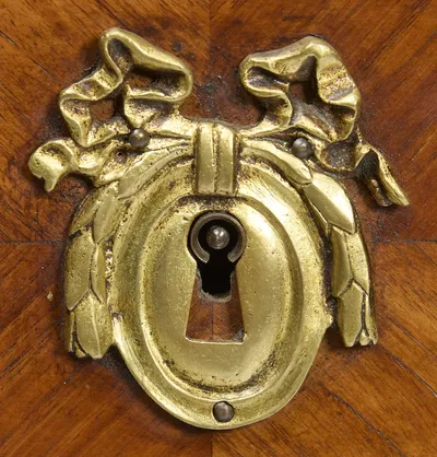 The gilt-metal keyhole surround cast as a ribbon bow with leafy pendants, set at the meeting of the quartered veneers between beaded brass borders.